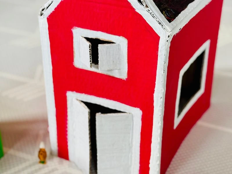 elbstgebasteltes Bauernhaus aus Karton mit roter Fassade, weißen Fensterrahmen und geöffneten Fensterläden.