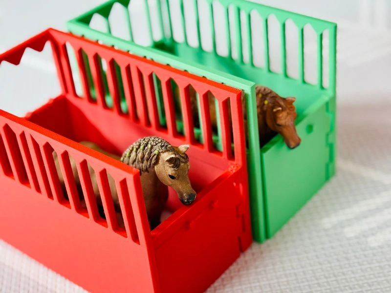 Zwei Pferdefiguren stehen in bunten, 3D-gedruckten Stallboxen in Rot und Grün mit detaillierten Gitterelementen.