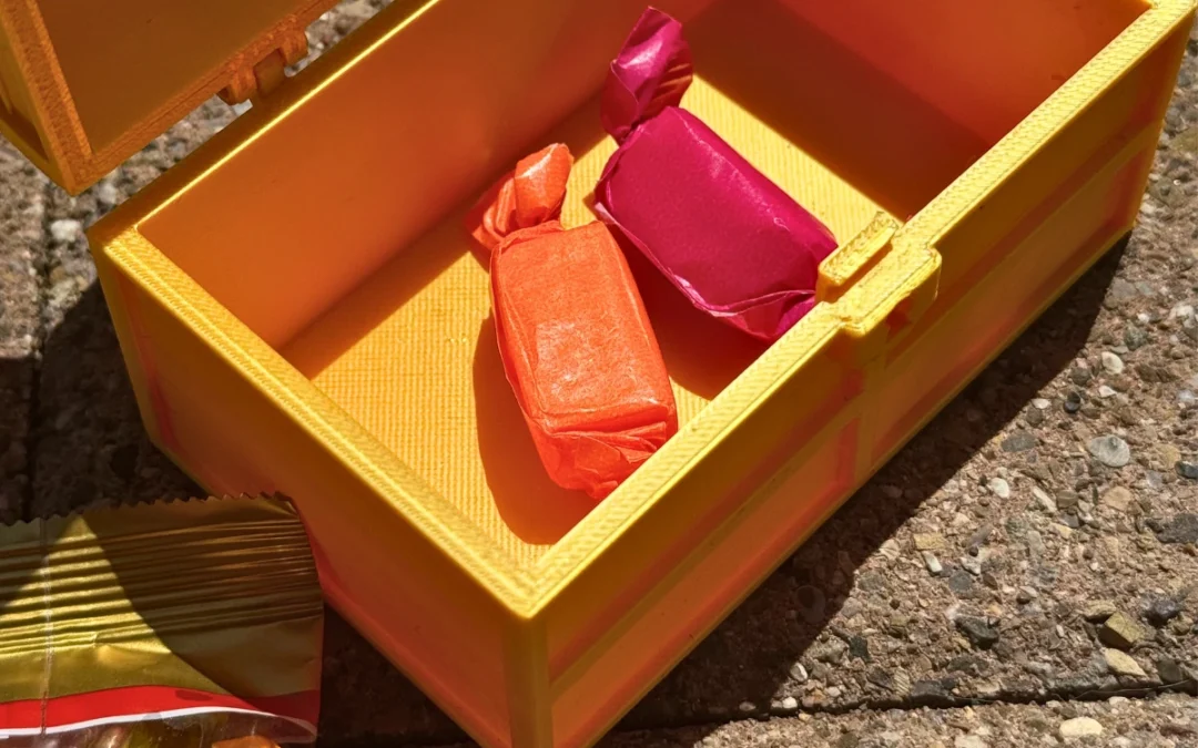Gelbe 3D-gedruckte Schatztruhe mit zwei farbenfrohen Bonbons im Inneren, liegend auf Steinboden im Sonnenlicht.