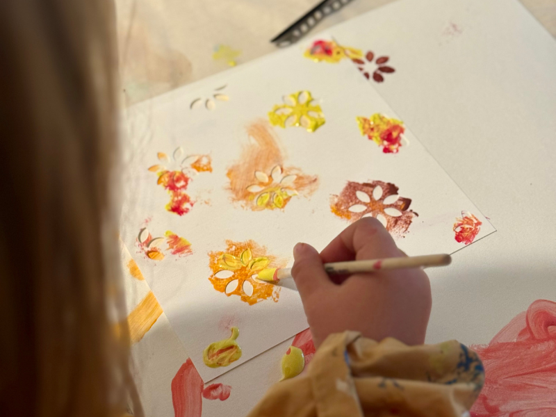 Kinderhand legt Buntstift in grünen 3D-gedruckten Dino-Stifthalter auf hellem Holztisch, umgeben von farbigen Stiften und Kinderzeichnung – kreative Geschenkidee aus dem Adventskalender 3D-Druck Teil 2 von BuildPlay3D.