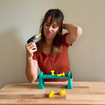 3D-Druck Fail oder Meisterwerk? – Mama testet Kinderwerkbank Frau sitzt vor einer 3D-gedruckten Kinderwerkbank und blickt skeptisch auf den Akkuschrauber – humorvolle Szene rund um den 3D-Druck-Test eines DIY-Spielzeugs für Kinder.