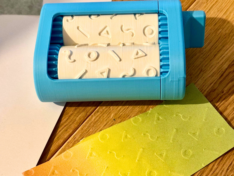 3D-gedruckte Prägewalze in Blau mit Mustern, die Strukturen auf buntes Papier prägt – ideal zum Basteln von Karten und DIY-Projekten.