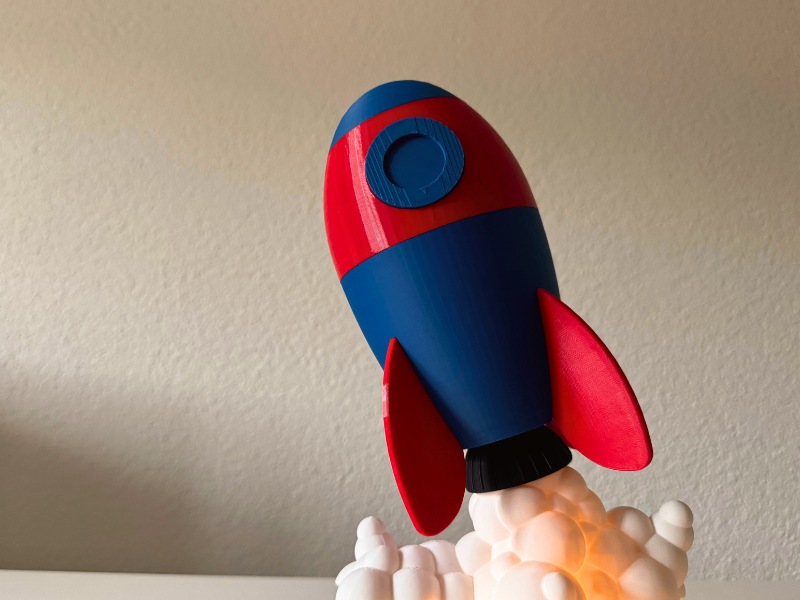 Raketen-Spardose mit Name 3D-Printing 3D-gedruckte Raketen-Spardose in Blau und Rot, aufsteigend aus weißen Wolken mit Licht darunter, auf einem hellen Untergrund vor neutraler Wand.