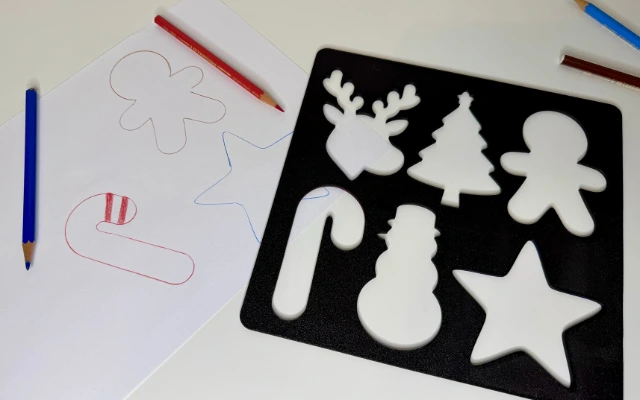 3D-gedruckte Weihnachtsschablone mit Motiven wie Stern, Schneemann und Tannenbaum auf Tisch mit Farbstiften