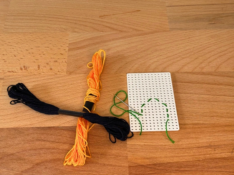 Stickvorbereitung auf Holzuntergrund – Stickplatte mit grünem Faden, Nadel und Garn in Orange und Schwarz.
