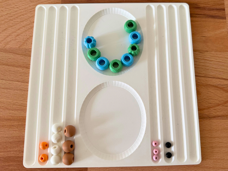 3D-gedrucktes Perlentablett mit bunten Bastelperlen in verschiedenen Farben, angeordnet in einem Oval, auf einem Holztisch.