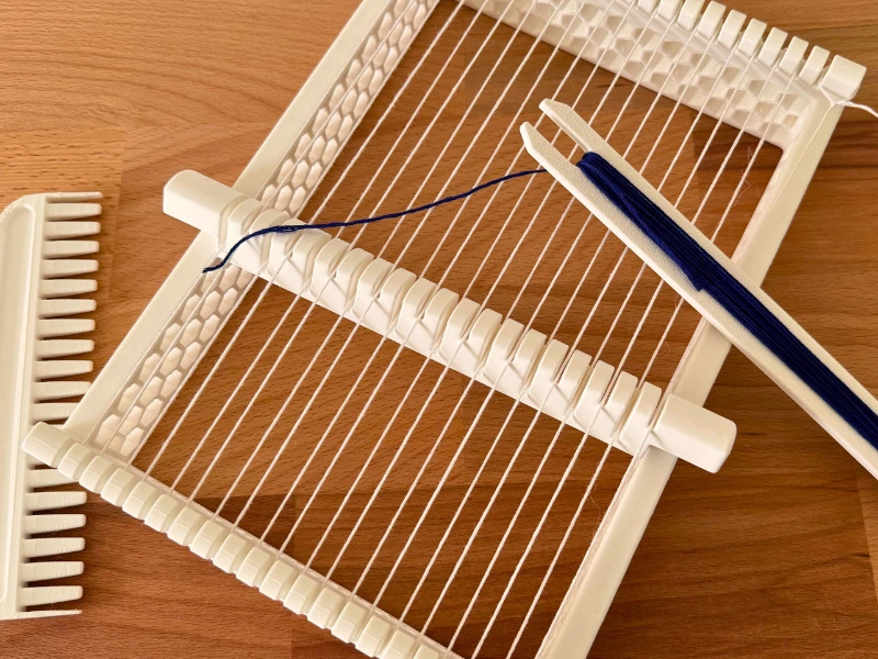 3D-gedruckte Webrahmen 🖼️ Alt-Text 3D-gedruckter Kinder-Webrahmen mit eingefädeltem blauen Garn auf einem Holztisch, bereit zum Weben.