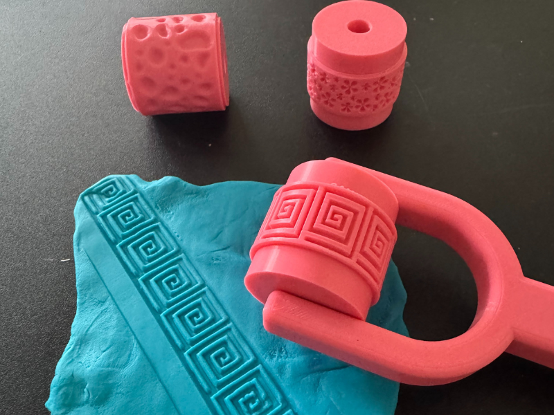 3D-gedruckte Texturwalzen in Pink prägen Muster in blaue Knete auf einem Tisch.