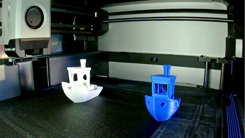 Zwei 3D-gedruckte Benchy-Boote, eines weiss und eines blau, stehen auf dem Druckbett eines Bambu Lab 3D-Druckers und zeigen unterschiedliche Druckqualitäten.