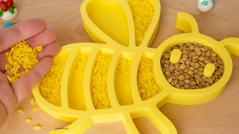 Bienen-Sensoriktablett von oben, gefüllt mit gelbem Reis und braunen Linsen, Hand hält gelbe Reiskörner, 3D-gedrucktes Montessori-Spielzeug