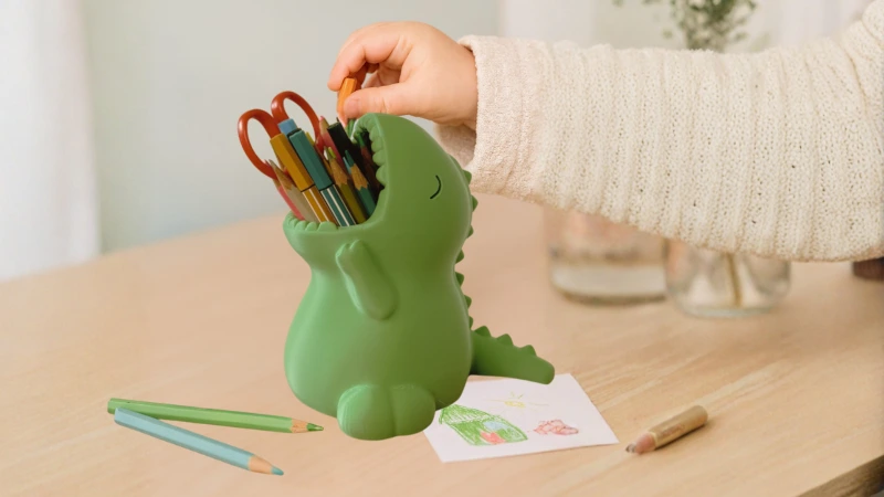 Kinderhand legt Buntstift in grünen 3D-gedruckten Dino-Stifthalter auf hellem Holztisch, umgeben von farbigen Stiften und Kinderzeichnung – kreative Geschenkidee aus dem Adventskalender 3D-Druck Teil 2 von BuildPlay3D.