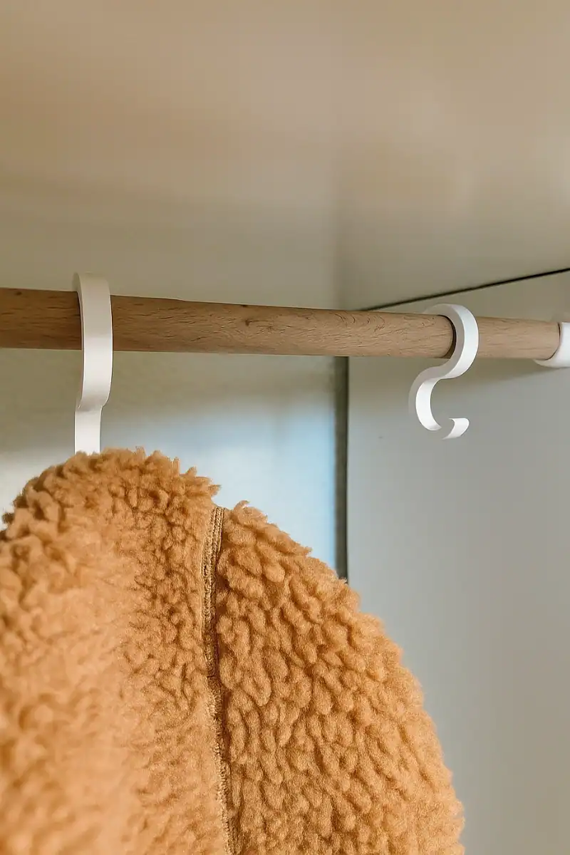 Nahaufnahme zweier 3D-gedruckter, weißer S-Haken an einer Holzstange im IKEA-Kallax-Regal, an denen eine Kinderjacke hängt.