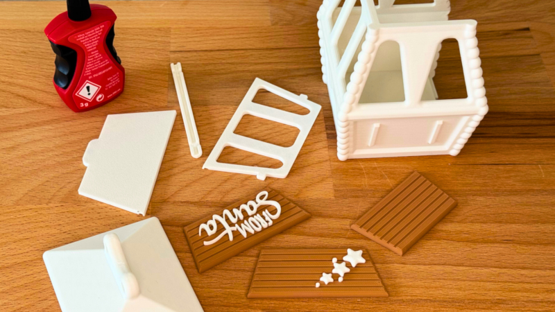 3D-gedruckte Gondel-Teile aus weißem und braunem PLA werden auf einer Holzoberfläche vorbereitet – inklusive Schriftzug „From Santa“ und Sekundenkleber.