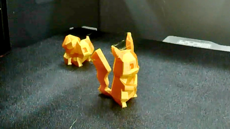 3D-Druck Problem: Stringing zwischen den Modellen Zwei orangefarbene, low-poly 3D-Figuren mit sichtbaren feinen Filamentfäden zwischen einzelnen Bereichen, ein typisches Beispiel für Stringing beim 3D-Druck.