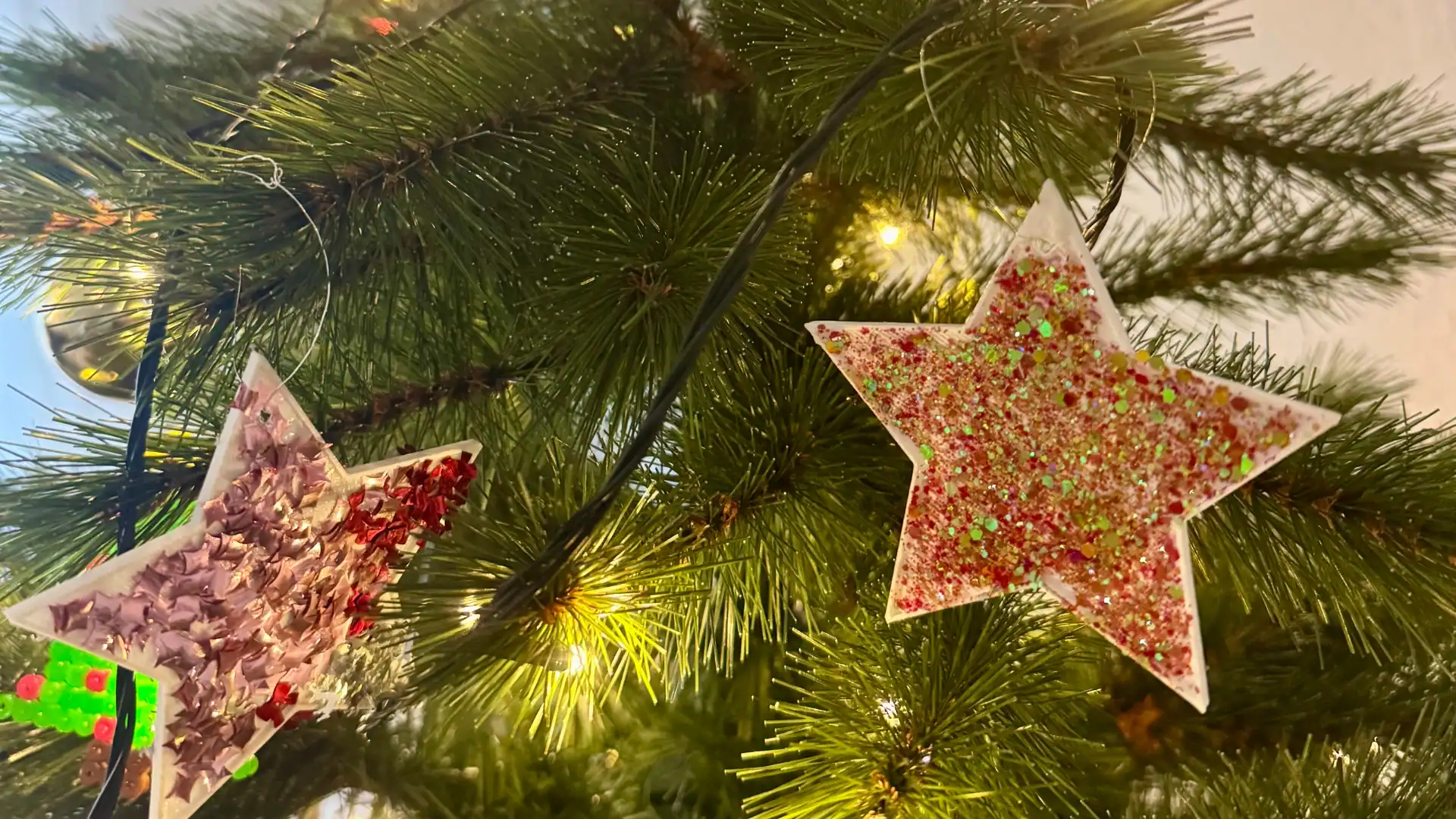 Zwei selbstgemachte Weihnachtssterne aus 3D-gedruckten Schablonen hängen geschmückt mit Glitzer am Weihnachtsbaum. Lichterkette und Tannenzweige schaffen eine festliche Stimmung.