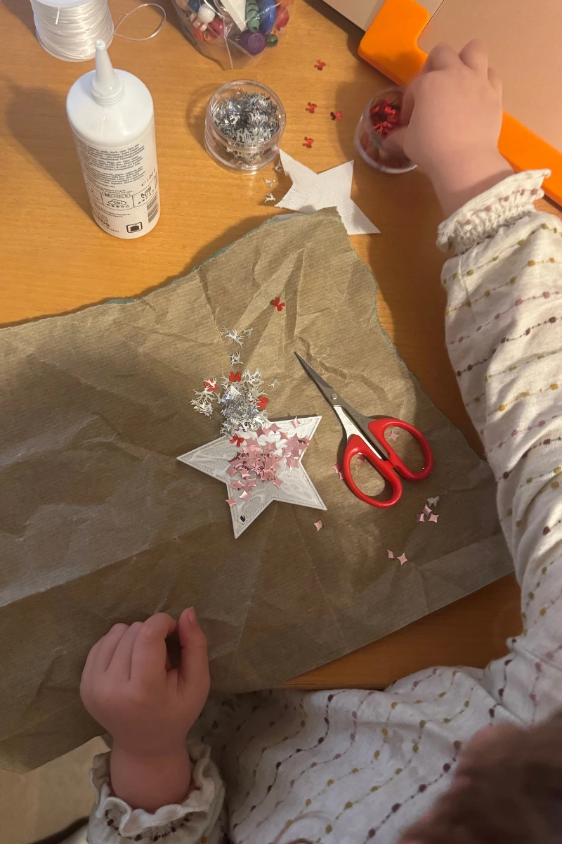 Ein Kind dekoriert einen weißen 3D-gedruckten Stern mit Glitzer und ausgestanzten Formen. Auf dem Tisch liegen Bastelschere, Leim und weihnachtliche Streudeko.