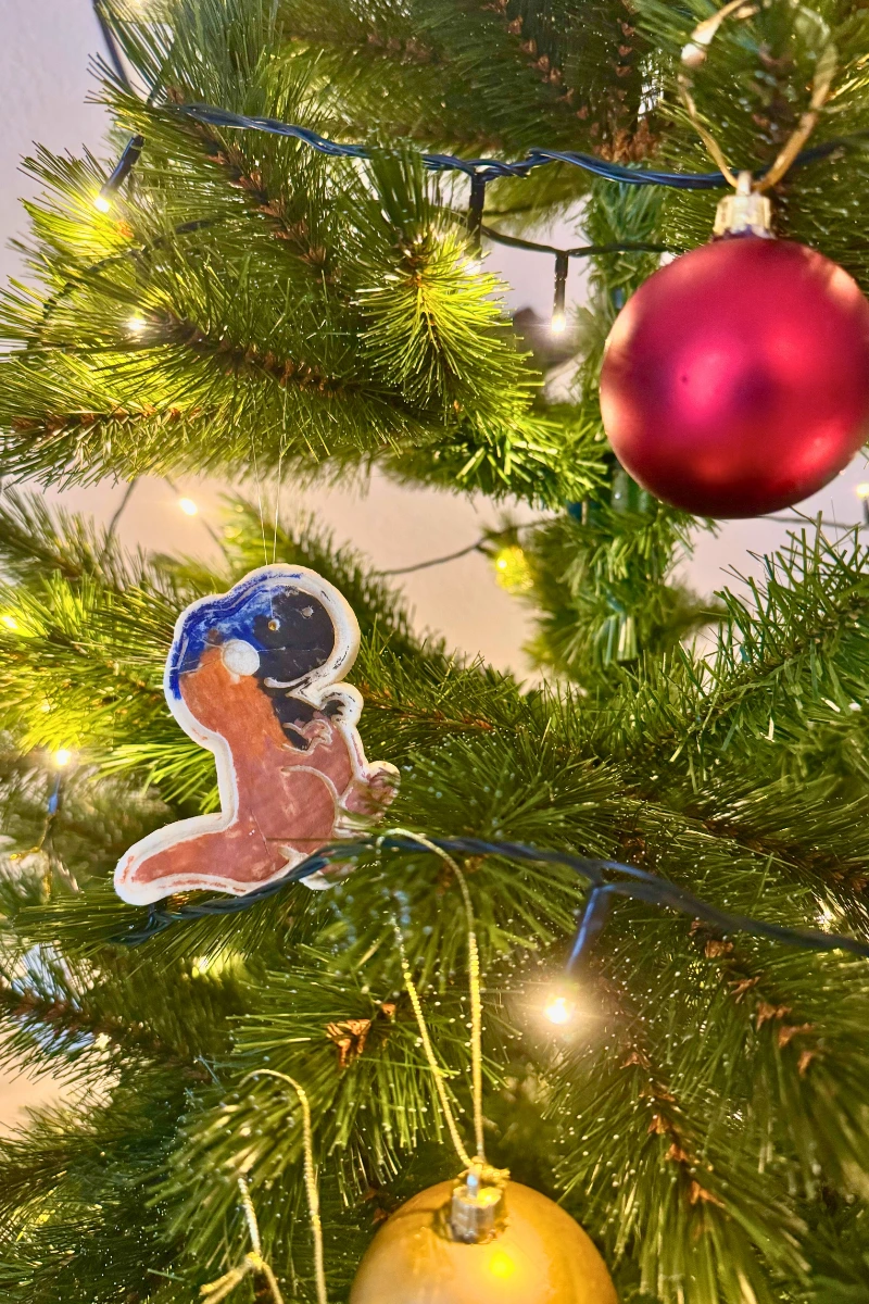 Salzteig-Weihnachtsanhänger Handgemachter Dino-Weihnachtsanhänger aus bemaltem Salzteig hängt zwischen grünen Tannenzweigen am Weihnachtsbaum. Im Hintergrund leuchten Christmas-Lichter und eine rote Weihnachtskugel.