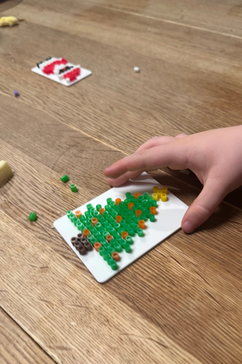 Weihnachtsschmuck aus Bügelperlen Ein Kind legt bunte Bügelperlen auf ein weißes 3D-gedrucktes Bügelboard, um einen Weihnachtsbaum zu gestalten. Einzelne Perlen liegen auf dem Holztisch, im Hintergrund ist ein weiteres weihnachtliches Bügelbild zu sehen.